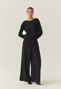 Schwarzer Pullover mit Rautenmuster und Verzierungen, kombiniert mit hoch taillierten, weiten schwarzen Hosen und spitzen schwarzen Absätzen.