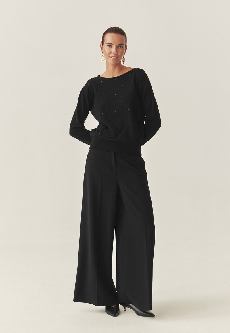 Schwarzer Pullover mit Rautenmuster und Verzierungen, kombiniert mit hoch taillierten, weiten schwarzen Hosen und spitzen schwarzen Absätzen.