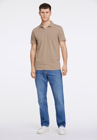 Polo shirt corto beige con colletto classico, abbinato a jeans blu a gamba dritta e sneakers bianche su uno sfondo neutro.