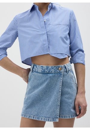 Mujer que lleva una camisa corta de rayas azules y blancas con mangas remangadas y una falda cruzada de mezclilla azul claro con cierre de botones.