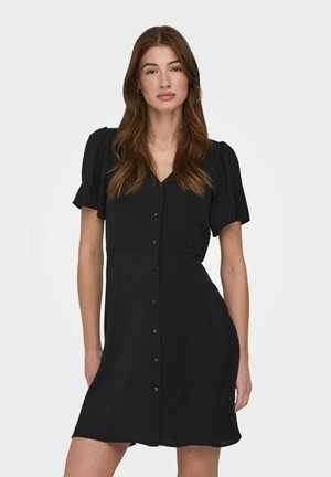 Kvinne med langt brunt hår iført en kort svart kjole med knapper og puffete korte ermer mot en ensfarget hvit bakgrunn.