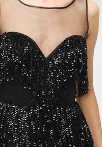Black dress featuring a sheer, sleeveless design with a deep V neckline. The bodice is adorned with cascading black sequins.