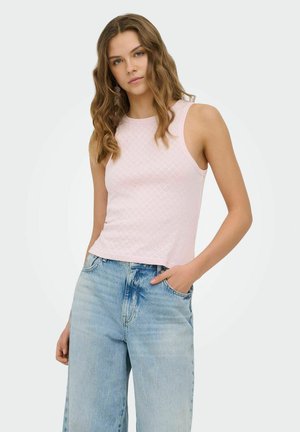 Woman with wavy hair wears sleeveless light pink textured top and light blue high-waisted jeans, standing with one hand in pocket.