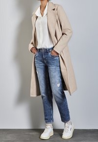 Beige ullblandad lång kappa, vit skjorta med knappar, slitna blå jeans och vita sneakers med gröna detaljer, presenterad mot en neutral bakgrund.