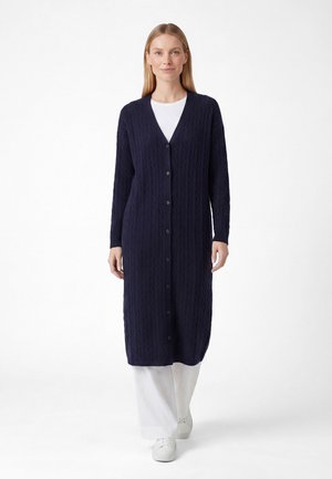 Vrouw draagt een lange navyblauwe kabelgebreide cardigan met knopen over een witte top, witte broek en witte sneakers, staand tegen een witte achtergrond.