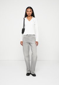 Donna in piedi con una maglietta bianca a maniche lunghe, jeans grigi a gamba dritta, scarpe nere e una borsa a tracolla nera su uno sfondo bianco.