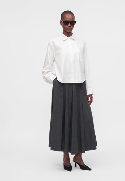Femme portant des lunettes de soleil noires, une chemise blanche boutonnée, une longue jupe plissée gris foncé et des talons noirs à bout pointu.
