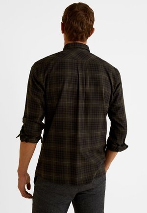 Mann, der ein dunkelgrünes und schwarzes kariertes Hemd mit hochgekrempelten Ärmeln trägt, von hinten vor weißem Hintergrund gezeigt.