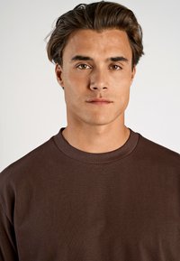 Brun långärmad skjorta med rund halsringning, som har en mjuk textur och ett subtilt ribbat tygmönster. Modellen har kort hår.