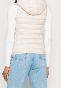 Gilet imbottito beige con cappuccio, caratterizzato da linee trapuntate orizzontali, abbinato a una maglietta bianca a maniche lunghe a coste e jeans blu chiaro.