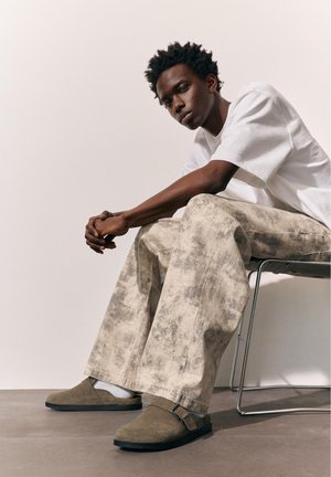 Junger Mann sitzt auf einem Metallstuhl, trägt ein weißes T-Shirt, beige gemusterte weite Hose, weiße Socken und braune Slipper.