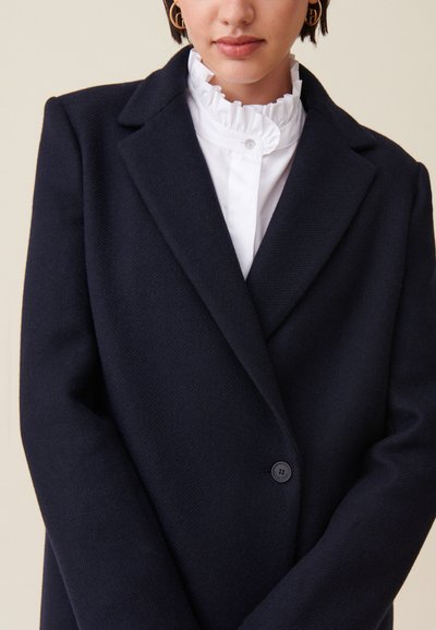 Cappotto in misto lana blu navy con design sartoriale, revers a punta, chiusura con un bottone e superficie testurizzata. Sotto indossa una camicetta bianca con volant.