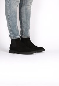 Bottines en daim noir avec des panneaux élastiques sur les côtés et une semelle en caoutchouc plate, positionnées à côté d'un jean en denim bleu clair, retroussé aux chevilles.