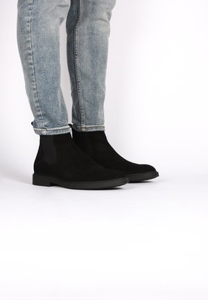 Botines de ante negros con paneles laterales elásticos y suela de goma plana, colocados junto a unos vaqueros de denim azul claro, remangados en los puños.