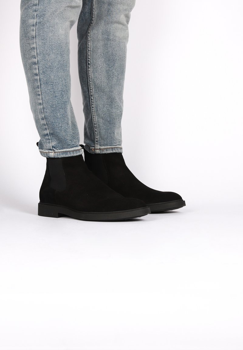 Bottines en daim noir avec des panneaux élastiques sur les côtés et une semelle en caoutchouc plate, positionnées à côté d'un jean en denim bleu clair, retroussé aux chevilles.