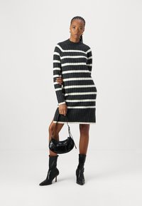 Robe pull tricotée avec des rayures horizontales noires et crème, col rond, manches longues et texture côtelée. Portée avec des bottes à talons noires et un sac à main.