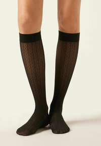 Piernas con medias negras transparentes de altura hasta la rodilla con un diseño geométrico de diamantes y rayas, de pie con los pies cruzados sobre un fondo claro.