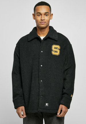 Chaqueta de sherpa negra con collar, cierre de botón y un logo bordado en amarillo con la letra "S" en el lado izquierdo del pecho. Puños acanalados y tejido texturizado.