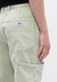 Ανοιχτοπράσινο παντελόνι cargo με πίσω τσέπη και ετικέτα με εμπορικό σήμα. Η υφή του υφάσματος φαίνεται λεία με ορατές λεπτομέρειες ραφής.