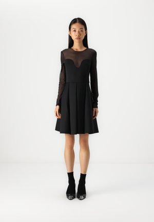 Vestito nero con maniche lunghe trasparenti, corpetto aderente e gonna svasata. Nota scollatura profonda e texture del tessuto liscia. Tacchi a stiletto neri.