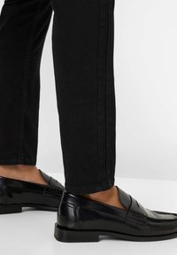 Mocassins en cuir noir avec une finition brillante, présentant un design cousu et un talon bas, associés à un pantalon noir slim fit.