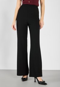 Wide-leg black pants with a textured fabric, high waistband, and smooth finish, paired with black pointed-toe heels featuring bow accents.