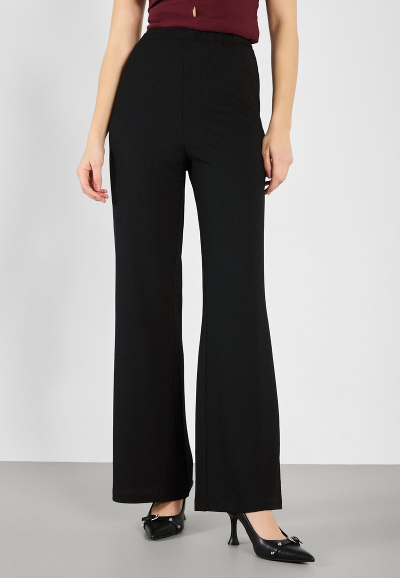 Wide-leg black pants with a textured fabric, high waistband, and smooth finish, paired with black pointed-toe heels featuring bow accents.