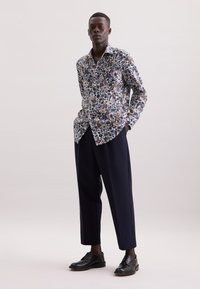 Camicia floreale a maniche lunghe nei toni di blu, marrone e verde, abbinata a pantaloni larghi scuri e scarpe in pelle nera. La camicia presenta un colletto a punta.