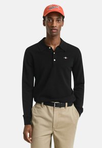 Zwart langslavend poloshirt met een kraag en geborduurd logo, gecombineerd met beige broeken en een roestkleurige pet met witte tekst.