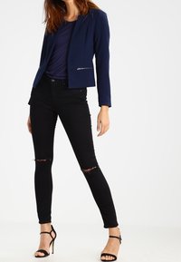 Námořnický přiléhavý blazer s kapsami na zip, kombinovaný s černými roztrhanými skinny džíny a černými sandály na vysokém podpatku. Jednoduchý, elegantní design.