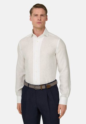 Man draagt een witte blouse met knopen, een riem met patroon en donkerblauwe broek, staand tegen een effen achtergrond.