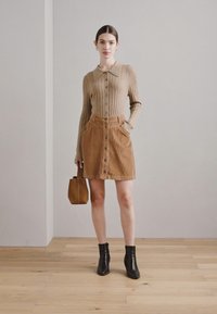 Gebreide beige cardigan met kraag en knoopsluiting, gecombineerd met een bruine corduroy rok met knopen aan de voorkant en zakken, zwarte enkellaarzen en een bruine handtas.