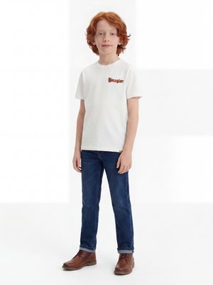 Αγόρι με κόκκινα μαλλιά στέκεται, φοράει λευκό μπλουζάκι Wrangler, μπλε τζιν και καφέ μπότες μπροστά σε απλό λευκό φόντο.