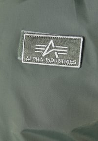 Olivgrön tyg med en rektangulär broderad applikation med logotypen "ALPHA INDUSTRIES" i ljusgrå och mörkgrå accenter.