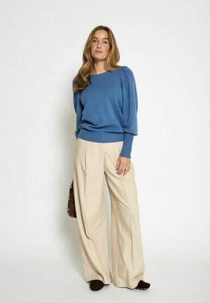 Pull en maille bleue à manches bouffantes, associé à un pantalon large beige clair. Le modèle tient un sac marron et porte des chaussures marron foncé.