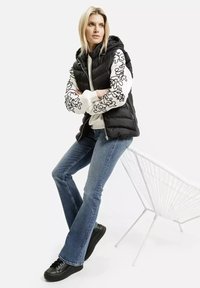 Czarna kamizelka puchowa z kapturem na białej bluzie z czarnymi abstrakcyjnymi wzorami. Niebieskie dzwony i czarne trampki, siedzący na krześle.