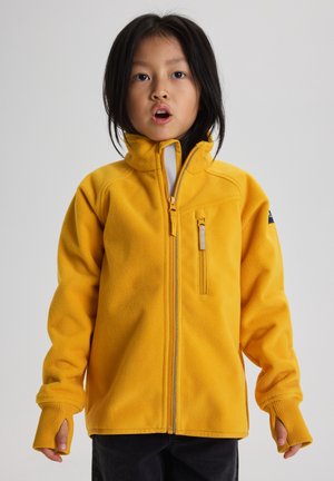 Kind draagt een gele fleecejas met hoge kraag en duimgaten, staand tegen een effen lichtgrijze achtergrond.