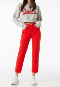 Sudadera gris corta con texto rojo "Santa Bárbara", combinada con jeans rojos de tiro alto y con pierna recta. Zapatillas blancas completan el conjunto.