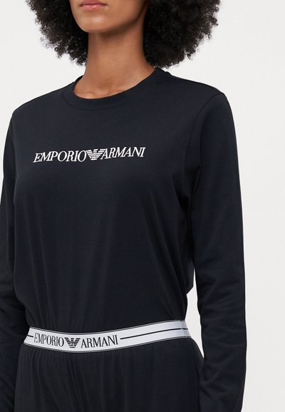 Melna krekliņa garām piedurknēm ar balto "EMPORIO ARMANI" logo, ar kontrastējošu balto logo joslu vidukļa zonā. Gluda tekstūra.