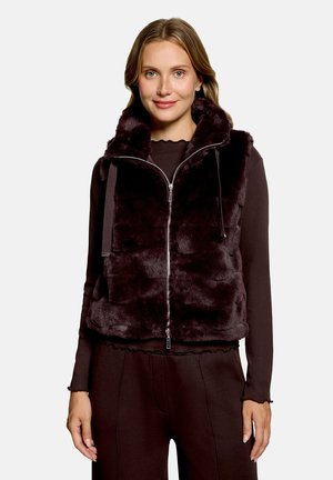 Femme portant un gilet zippé en fausse fourrure marron foncé sur un haut à manches longues et un pantalon assorti, debout devant un fond uni.