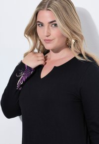 Haut noir à manches longues avec un décolleté en V et une broderie florale violette détaillée au niveau des poignets. Texture de tissu lisse.