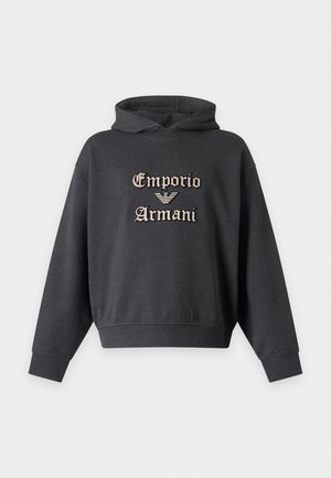 Νέος άνδρας στέκεται με τα χέρια στις τσέπες, φοράει σκούφο φούτερ με το λογότυπο Emporio Armani, φαρδιά σκούρα μπλε παντελόνια και μαύρα αθλητικά παπούτσια.