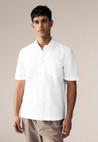 Majica kratkih rukava u bijeloj boji od laganog materijala s gumbima sprijeda i džepom na prsima. Glatka tekstura, krojena kroj, minimalistički dizajn.