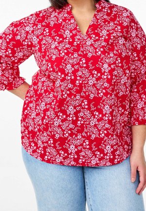 Frau trägt eine rote Bluse mit Blumenmuster und Dreiviertelärmeln sowie hellblaue Jeans, Hand ruht auf der Hüfte.