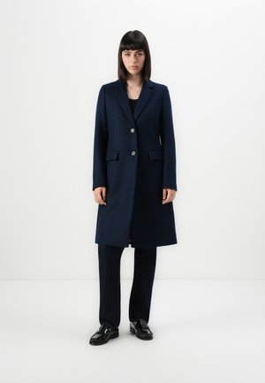 Donna in piedi con un cappotto blu navy lungo fino al ginocchio, pantaloni neri e mocassini neri su uno sfondo bianco.