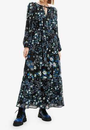 Kobieta ubrana w długą czarną sukienkę w kwiaty w kolorze niebieskim, białym i zielonym, połączoną z czarnymi i niebieskimi masywnymi botkami sznurowanymi.