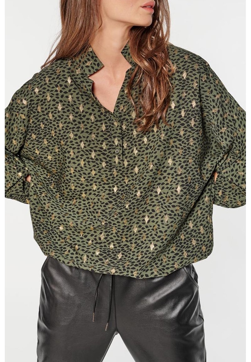 Femme portant un chemisier vert olive à motifs avec des accents métalliques et un pantalon en cuir noir, posant les mains sur les hanches.