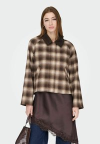 Kvinne iført en brun rutete jakke over et mørkt satengskjørt med blonder og blå jeans, holder en teksturert brun håndveske.