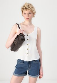 Hvit ermeløs topp med knappdetaljer, kombinert med blå denimshorts. Modellen holder en mørk brun lærveske. Glatt tekstur, uformell design.
