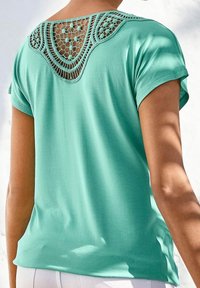 Teal t-paita lyhyillä hihoilla, jossa on koristeellinen virkkausmainen takasuunnittelu. Pehmeä materiaali ja rento malli, jossa on käännetyt hihat ja pyöristetty helma.